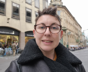Portrait of January Star Student Charlie Taylor smiling outdoors on a city street, wearing round glasses and a black shearling jacket, with historic buildings in the background.