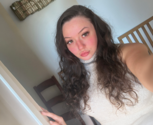 Young woman with long dark curly hair and a nose piercing wearing a sleeveless cream turtleneck top, taking a selfie indoors with natural light and a direct gaze toward the camera.