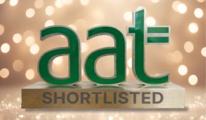 Here’s clear, accessible ALT text suitable for your website: ALT text: Green “aat” logo displayed on a gold-toned background with soft light bokeh effects, sitting above the word “SHORTLISTED” in bold capital letters.