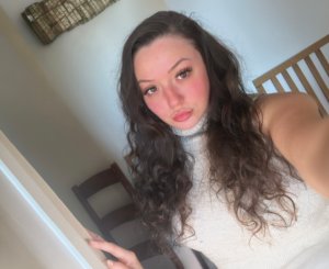 Young woman with long dark curly hair and a nose piercing wearing a sleeveless cream turtleneck top, taking a selfie indoors with natural light and a direct gaze toward the camera.