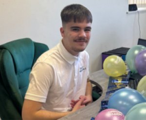 Alfie Weston, May Star Student at Accountancy Learning, smiling while seated at a desk decorated with colourful balloons in the office, wearing a white branded polo shirt.